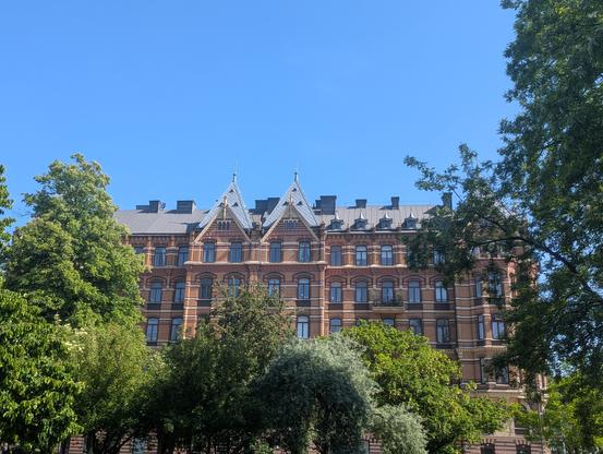 Belle façade d'un haut bâtiment en briques avec alternance de teintes. De nombreuses fenêtres reflètent le bleu du ciel. En bas, la façade est partiellement cachée par le feuillage des arbres.