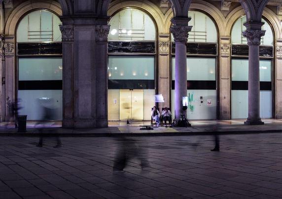A few people walks past the scene at night. We only see their motion blurs, while a couple sits under an arch of a building in the background, without being blurred. They look like they're surrounded by ghosts.