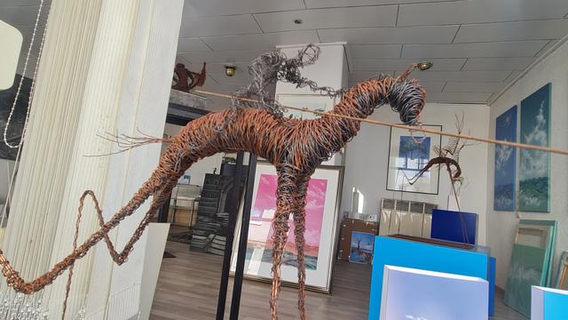 Blick durch Schaufenster in Raum, Bilder stehn und lehnen überall, auf podesten Figuren aus verwickeltem draht, stellen Reiter, Drachen, Engel und.ä. dar.