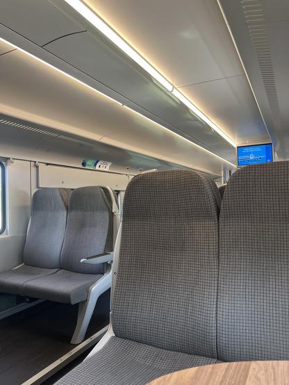 Innenraum eines modernen Zuges mit zwei leeren Sitzen mit grau gemusterten Polstern, einem Holztisch im Vordergrund, einer Deckenbeleuchtung und einer digitalen Anzeigetafel an der Wand.