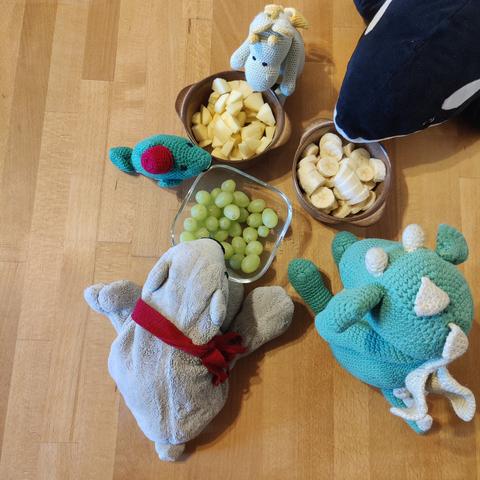 Eisbär Theo, Wal mit Hut, Orca Panda und die Drachen Arktos und Tabaluga sitzen un drei Schüsseln Obst herum. Jeweils eine Schüssel enthält Apfelstückchen, geschnittene Bananen bzw. Weintrauben.