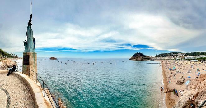 Color panorama with a large statue of the goddess Minerva holding a spear and mounted on a high seafront walkway overlooking the beach and town of Tossa de Mar