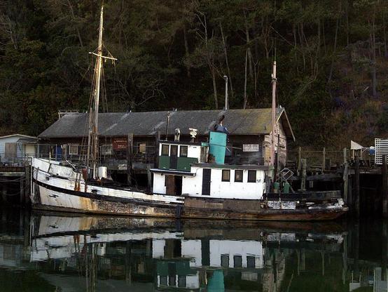 Working Boat

Tis is an image of an old fishing boat tied to a dock in Noyo Harbor in Ft. Bragg, California.
The foreground of the image consist of the still waters of Noyo Harbor. The water provides a near mirror finish reflecting the boat on the water.
The boat itself is in need of maintenance as can be seen. The side of the boat is missing most of it's paint. There is actually a plank missing from the bow.
Behind the boat is an old shack. The shack shares many characteristics as the boat does. IT needs paint and possibly a new roof.