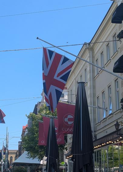 Façade d'un pub auquel est accroché un mât portant drapeau britannique. Derrière, on aperçoit un autre drapeau de couleur rose, portant l'inscription "CONSID". Mais comme il est partiellement caché par un parasol du pub, on croit lire "CONS".