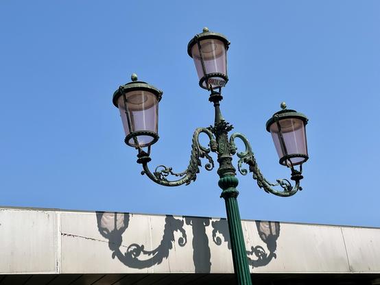 An ornate green wrought iron streetlight, with three arms topped with lanterns in  pink-tinged glass. The photo is taken looking up, so that most of the background is uninterrupted pale blue sky, but the lower fifth of the frame is a flat-topped white wall, and the lower bit of the streetlight - a bit of the pole, two curved scrollwork arms and the bottom edges do the bottom two lanterns - casts a clear black shadow against it.