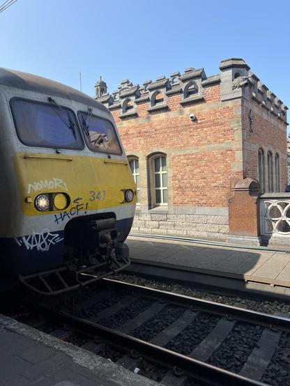 Ein Zug hält an einem Bahnhof, dessen Front links sichtbar ist. Im Hintergrund ist ein altes Gebäude aus roten Backsteinen mit Steinakzenten unter einem klaren blauen Himmel zu sehen. Graffiti ist im Zug vorhanden.