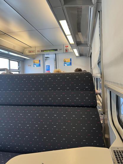 Innenansicht eines Zugabteils mit gemusterten Stoffsitzen, Deckenleuchten und einem Display mit Informationen.