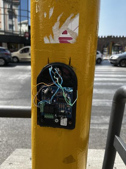 A close-up of a yellow traffic pole featuring exposed wiring and a circuit board attached. There is a partially visible sticker on the pole and vehicles in the background.