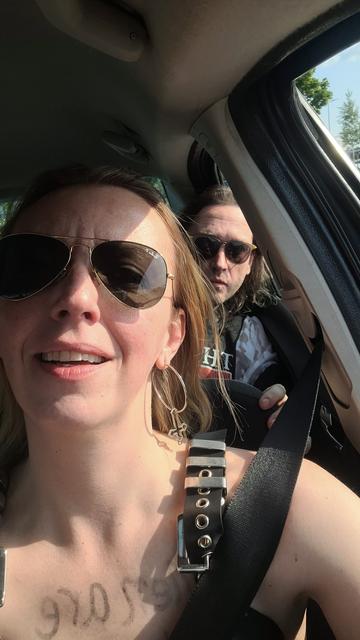 A woman in sunglasses takes a selfie inside a car, with another one is wearing sunglasses seated behind her. She has large cross-shaped upside down earring, a black top with metallic grommets, and a faint handwritten mark on her chest that appears to read "men at too emotional to lead." Sunlight is streaming through the car window, casting shadows on their faces.