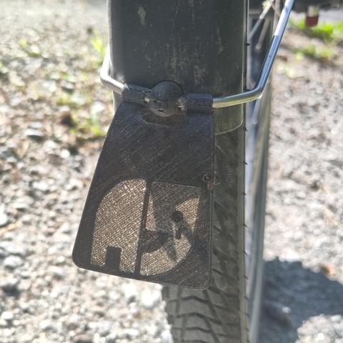 Fahrradvorderrad: Nahaufnahme von hinten, Reifen, unteres Ende des schwarzen Schutzblechs, silberne Haltestange, schwarzes Gummiknubbel als Halter, darunter ein schwarzer Spritzschutz, darauf durchscheinend der weinende Natenom-Elefant. Im Hintergrund ein Schotterweg