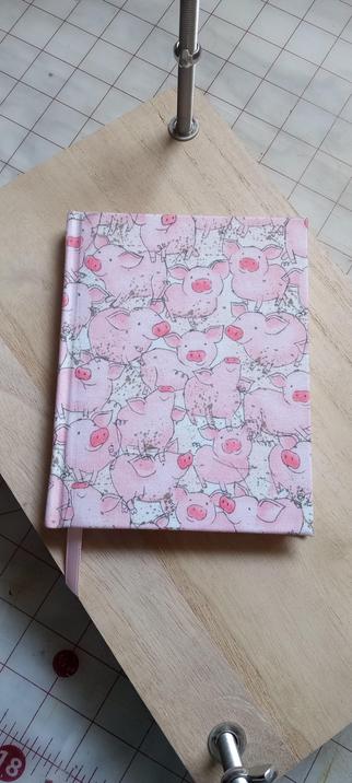 A 4,5" X 5.75" book on top of a book press, on a work bench. The book is covered in pink fabric with a drawing of pigs playing and wallowing in mud. The mud is depicted in little droplets. The pigs are round with floppy ears, curly tails and bright pink snouts and appear to be smiling. There is a pink ribbon extending from the binding of the book.