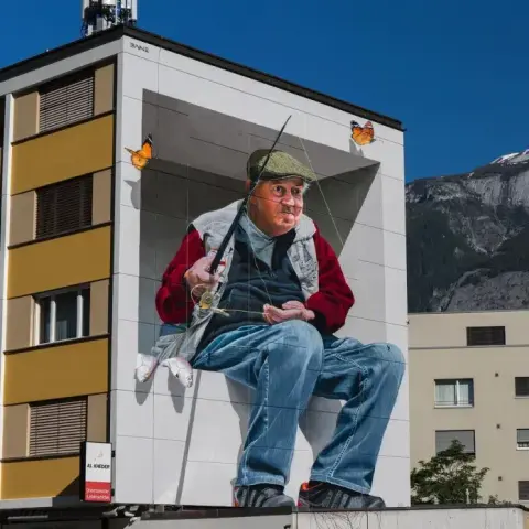 A stunning mural titled 'The Plessurfischer' by Fabian Bane Florin in Chur, Switzerland. The artwork features a lifelike portrayal of an elderly man in a fisherman's outfit, seated with a fishing rod. Monarch butterflies flutter around him, adding vibrancy. The mural is painted on the side of a multi-story building, with the backdrop of majestic mountains. Created for the Street Art Festival Chur, it captures a serene moment in nature with incredible detail and realism.
