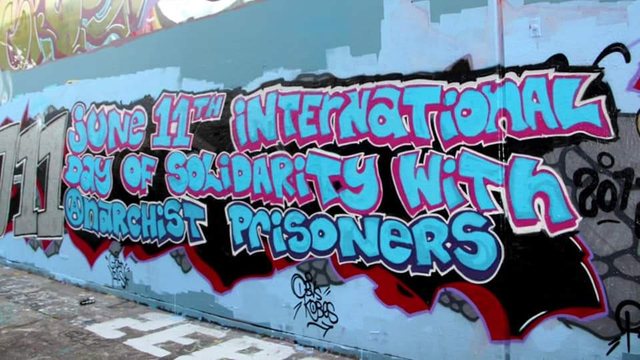 A photo of a graffitied wall that reads "June 11th International Day of Solidarity with Anarchist Prisoners" in blue bubble letters.
