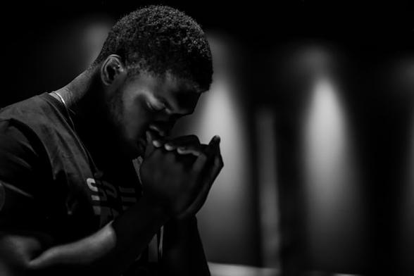 [A black-and-white photograph of a young Black man bowing his head in prayer, bringing his clasped hands to his mouth. He stands in a dimly lit space, possibly a house of worship.]