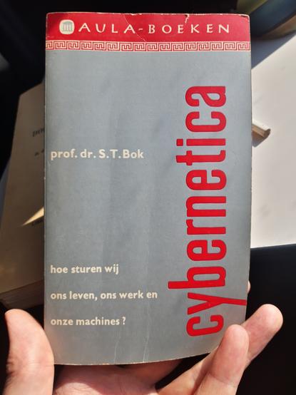 A hand holds a pocket book by prof.dr. S.T. Bok, entitled "Cybernetica, hoe sturen wij ons leven, ons werk en onze machines ? "