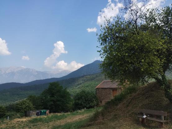 Wiese, Berge, Wolken: Links im Hintergrund der Gipfel Canigou, im Vordergrund links Mülltonnen, rechts eine Bank mit einem Buch drauf, im Hintergrund hellblauer Himmel mit Wolken.
