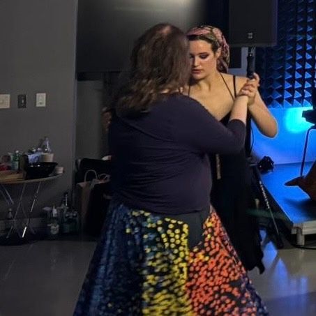 A woman’s rainbow-dotted skirt twirls as she pivots, led by a woman in a black dress and floral head scarf.