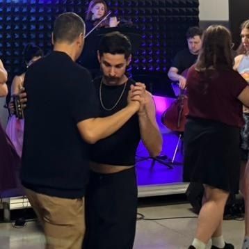 A mustachioed man in a black cropped tank top leads a man in a short sleeved shirt while another couple dances in the background and the band plays on stage.