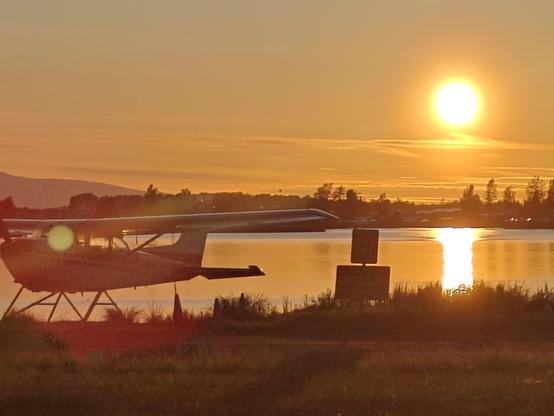 An orange-hued Sun, low on the horizon, turns an idyllic scene of a seaplane on a lake in Alaska sunset-colored, with a thin haze of clouds top-lit by the setting Sun's light.