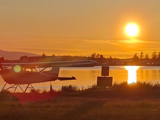 An orange hued Sun turns the landscape of a sea plane on a lake-like body of water into a sunset landscape, with thin, wispy clouds low on the horizon top-lit by the sunlight.