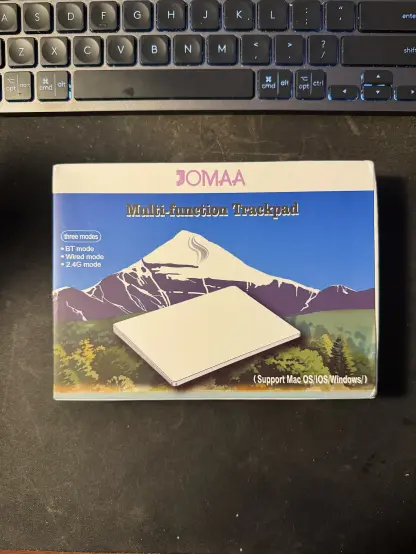 The outer box packaging of the JOMAA KB96 trackpad, showcasing the white trackpad with an art of a snowy mountain with a blue backdrop in the background. Written on the box that the trackpad supports three modes; BT, Wired, and 2.4G.