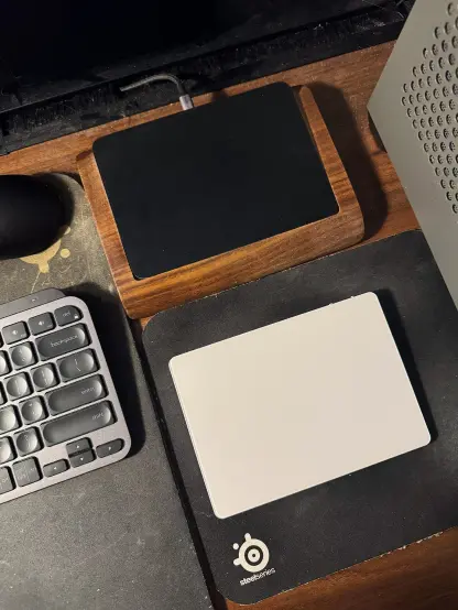 A topdown photo of the JOMAA KB96 trackpad in white, placed on a small black mousepad. On top of it is the Apple Magic Trackpad 2 in black, resting in a walnut, wooden tray. Next to them is a large, black mousepad with a Logitech MX Keys Mini keyboard in graphite.
