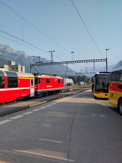 Foto. Ein roter Zug und zwei gelbe Busse im Bahnhof.