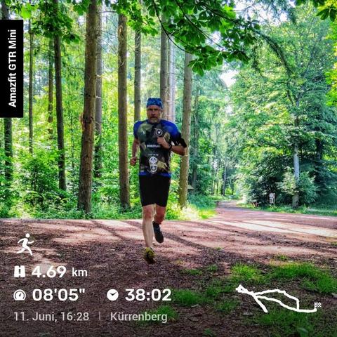 Ein Mann läuft auf einem Waldweg, umgeben von hohen Bäumen und grünem Laub. Er trägt ein blau-schwarzes Laufshirt mit verschiedenen Logos und schwarze Shorts. Links im Bild steht 'Amazfit GTR Mini'. Unten sind Laufdaten eingeblendet: 4,69 Kilometer, 08'05" Pace, 38:02 Minuten Laufzeit. Darunter stehen Datum, Uhrzeit und Ort: 11. Juni, 16:28, Kürrenberg. Rechts unten zeigt eine kleine Karte die Laufstrecke.