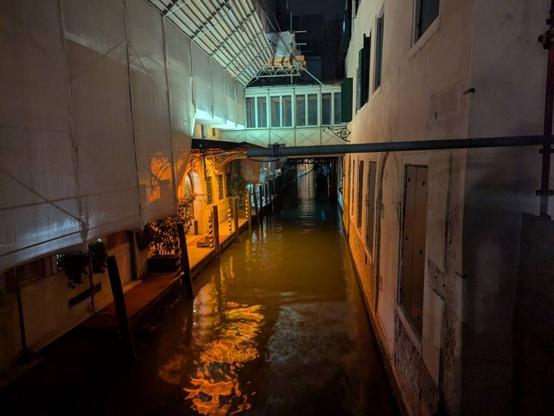 A dark canal in Venice with an orange light entrance right off the water.  The water is calm giving off interesting reflections of the light.  The upper walls of the canal are white and there is some sort of walking structure in the background crossing the water.