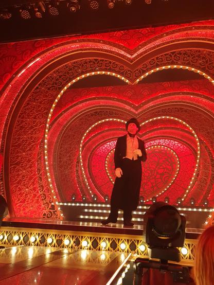 Stage of the Moulin Rouge musical, consisting of red heart shapes decorated with lights and frills. In front of the stage, a small part of a catwalk leading towards the audience is visible.

A man in a white tie suit, holding a sigarette is standing center stage.
