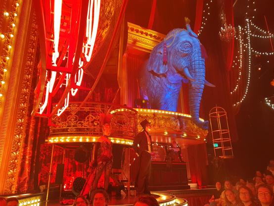 Side stage of the Moulin Rouge musical, consisting of small bar, with a blue elephant on its roof. To the left is a small balcony where people could sit, to the right is a cage-like structure where someone could stand and dance in.

In front of the bar is a man in white tie suit and a woman in burlesque outfit.

Overall coloring is red and gold, with lots of small decorations and lights.