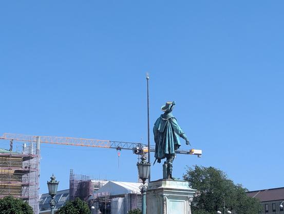 Statue d'un homme debout, de dos, l'index de la main droite pointé vers le sol. La statue est située sur un piédestal assez élevé. En arrière-plan se trouvent divers bâtiments et une grue de chantier. La photo a été prise sous un angle qui donne l'impression que l'homme appuie, de son doigt tendu, au bout de la grue.