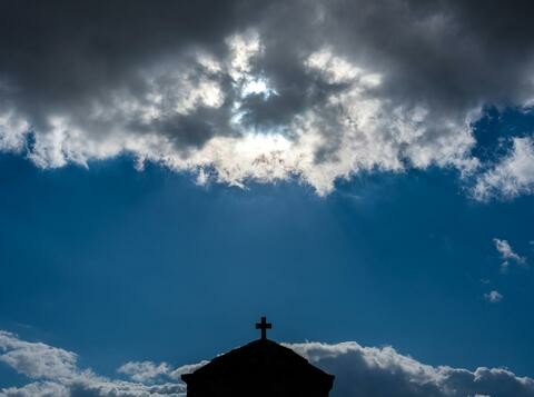 A blue sky with the sun behind a cloud. A silhouette of a cross and building visible at the bottom.