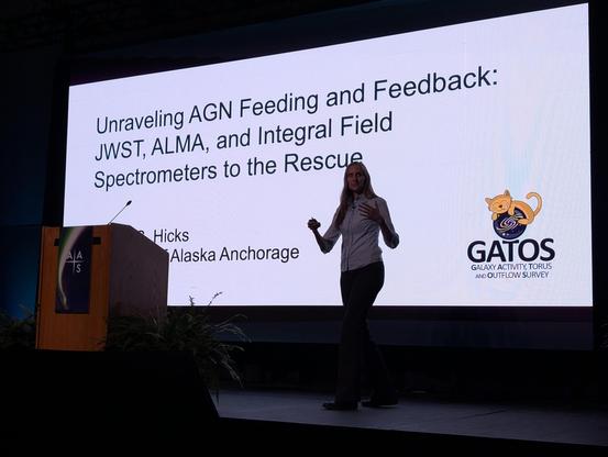 Erin Hicks stand on stage in front of a bright screen showing slides.
Unraveling AGN Feeding and Feedback: JWST, ALMA, and Integral Field Spectrometers to the Rescue, Erin Hicks, University of Alaska Anchorage.
Logo for GATOS: Galaxy Activity, Torus and Outflows Survey, a cute cat embracing a spiral galaxy.