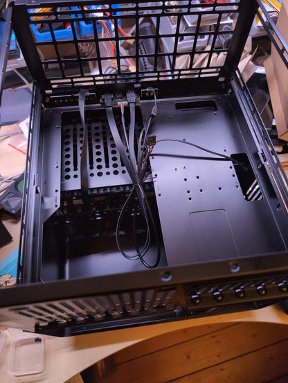 Das Bild zeigt den Innenraum einer leeren Computerbox, die auf einem Holztisch steht. Die Box ist aus schwarzem Metall und hat eine offene Frontplatte mit einem Gitterdesign, das zur Belüftung dient. Im Inneren sind mehrere Lüftungslöcher und Halterungen für Hardwarekomponenten zu sehen. Einige Kabel, darunter eine breite Steckdose-Kabel und mehrere dünnere Kabel, sind an den Rücken der Box angeschlossen. Die Kabel sind ordentlich angeordnet und verlaufen entlang der Innenseite der Box. Im Hintergrund sind weitere elektronische Komponenten und Bauteile sichtbar, die auf einem Tisch liegen. Die Umgebung deutet auf eine Werkstatt oder einen Bastelraum hin.
By @altbot