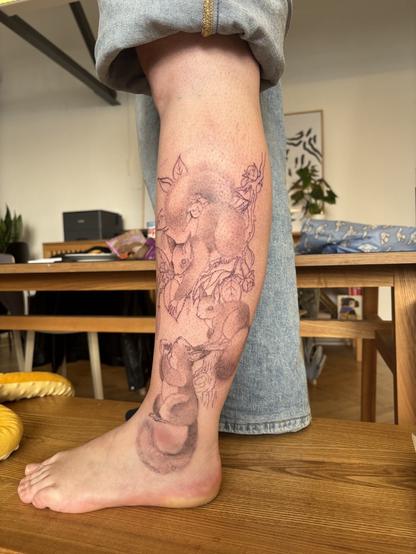 Bild von meinem linken Bein. Ich trage eine Jeans, die ich hoch gekrempelt habe.
Auf dem bei ist ein frisches Tattoo:
Drei Eichhörnchen. Eines hat eine Nuss in den Pfoten, die es nach oben streckt. Eins sitzt auf einem Ast und versucht die Nuss von oben zu greifen. Ein drittes Eichhörnchen sitzt auf einem Baumstumpf und beobachtet das ganze.
Die „Lücken“ sind mit Ästen und Blättern eines Haselnussbaums gefüllt.
Alles ist in schwarz tönen.
