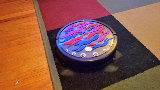 A circular robot vacuum with six googly eyes of various sizes stuck on the front and tiger stripes are literally pink, red and blue foam across the body. The brushes in the front look like little crab mouth parts