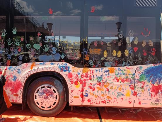 Foto vom hinteren Teil eines Linienbus, dieser, in der Grundfarbe weiß, wurde durch viele Kinderhände bunt bemalt. Kinderhandabdrücke, Vornamen, Zickzacklinien, etc. Am linken Bildrand sieht man eine vollständig rot gefärbte Kinderhand, die auf eine Malerei auf dem Bus zeigt.