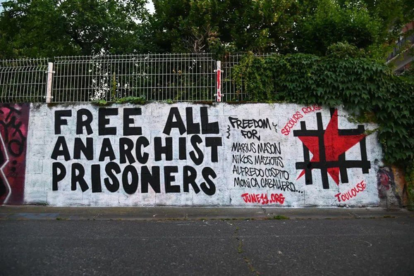 Graffiti reading "Free all anarchist prisoners—freedom for Marius Mason, Nikos Maziotis, Alfedo Cospito, Monica Caballero... june11.org"