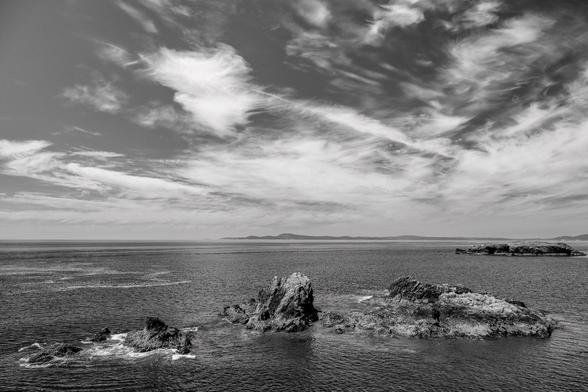 Urchin Rocks, a collection of three small rocky islands near Rosario Head in Deception Pass State Park, left to right across the bottom of the frame.  White waves are washing against the leftmost rock.  In the middle distance, on the left, there is a rocky island.  Above, the sky is full of wispy clouds.