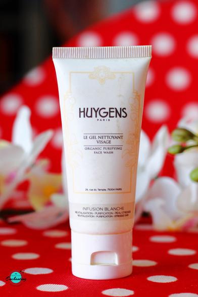 A close-up image of the Huygens Paris Infusion Blanche Organic Purifying Face Wash tube, placed against a red polka dot background with soft floral decorations in the frame. Full Review is featured on the blog 'Natural Beauty And Makeup'.