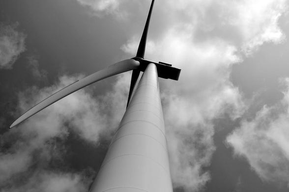 View upwards directly under the rotor blade of a wind turbine. Due to the proximity of the camera to the tower, it appears very important and leads optically very high into the sky. One of the three rotor blades can be seen in its entirety and complements the tower in the image composition. Some clouds can be seen in the sky. The image is in black and white