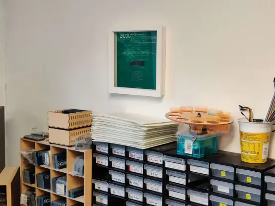 View of the side wall of the office which is pained white. At the bottom of the shot are component storage drawers and pigeon hole PCB storage with some things piled on top. Above that is a green PCB in a white box frame. In the centre is a black 40 pin chip and the word Zilog on the silkscreen of the PCB with some other information also on it.