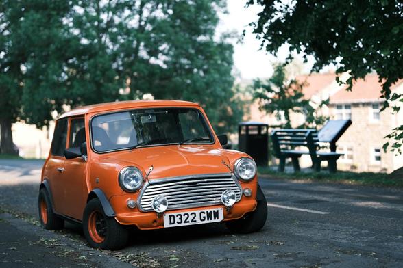 A old, small, orange car (Mini) is parked on the side of the road, its wheels turned as if to drive off. The bright orange of the car contrasts with the muted greens of the surrounding trees and the gray of the road.
