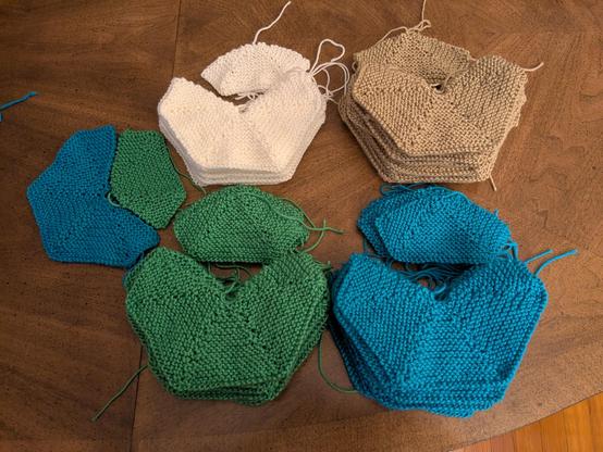 Piles of knitted shapes, some large and some small, on a wooden table. The piles are arranged by colour: air-freshener green, sky blue, sand tan, and lane-divider white. On the left, a large blue and a small green shape are shown to fit together to form a hexagon.