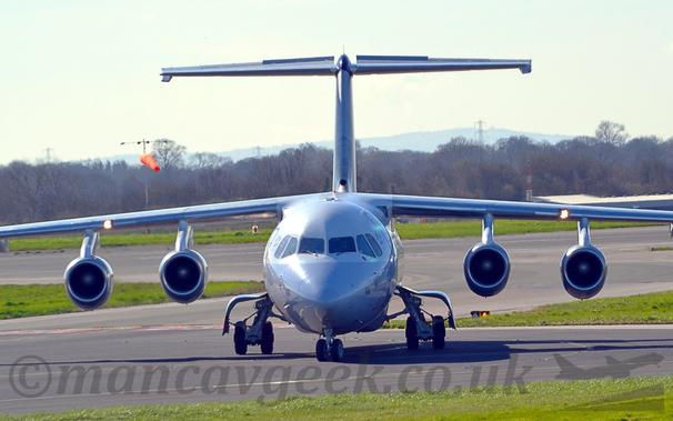 Head-on view of a high-winged, 4 engined jet airliner taxiing towards the camera, it's nose-wheel turned slightly to it's right.
The plane has white upper surfaces, and a grey lower half.
Green grass lines the taxiway in the foreground, and the runway in the background, leading up to trees in the distance, under a bright but hazy sky.