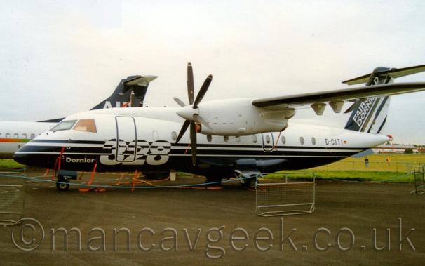 Side view of a high-winged, twin propellor-engined airliner parked on black tarmac facing to the left.
The plane has white upper surfaces, with dark blue lower surfaces, and a large white number "328" on the lower forward fuselage.
The black registration D-CITI is on the upper rear fuselage, under the dark blue and white tail, which has white "Fairchild Dornier"  at the front.
Sections of metal fence joined with long pieces of blue rope are placed in front of the plane.
The rear fuselage and tail of another airliner can be seen on the left, with grey sky filling the rest of the frame.
