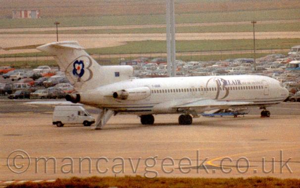 Rear shot of a 3 engined jet airliner with the engines mounted on the rear fuselage.
There are blue "BelAir" titles on the upper forward fuselage, overlaid with a large grey letter "B".
A similar letter "B" is on the tail, next to a dark blue, white, and red stylised flower.
The black registration "F-GGGR" is on the upper rear fuselage, under the tail.
A set of built-in airstairs is coming out of the rear fuselage, under the tail, next to a white van.
Grey concrete apron fills the foreground, with parked cars and green grass leading off into the distance in the background.
