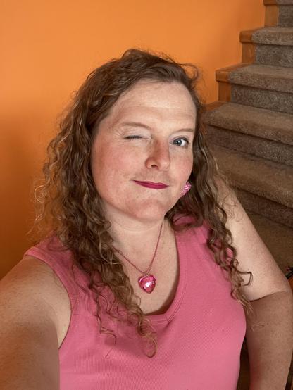 miriam, a white trans woman with long curly dirty blonde hair, wearing a pink ribbed tank, a chunky pink heart pendant, and sparkly pink heart earrings. she is winking at the camera.