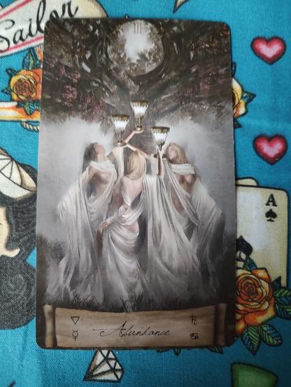 Card from the Heaven and Earth Tarot deck. 
Three of Cups.
Three women wearing white robes dancing and forming a circle. They are holding chalices (cups) up high, all under a tree.